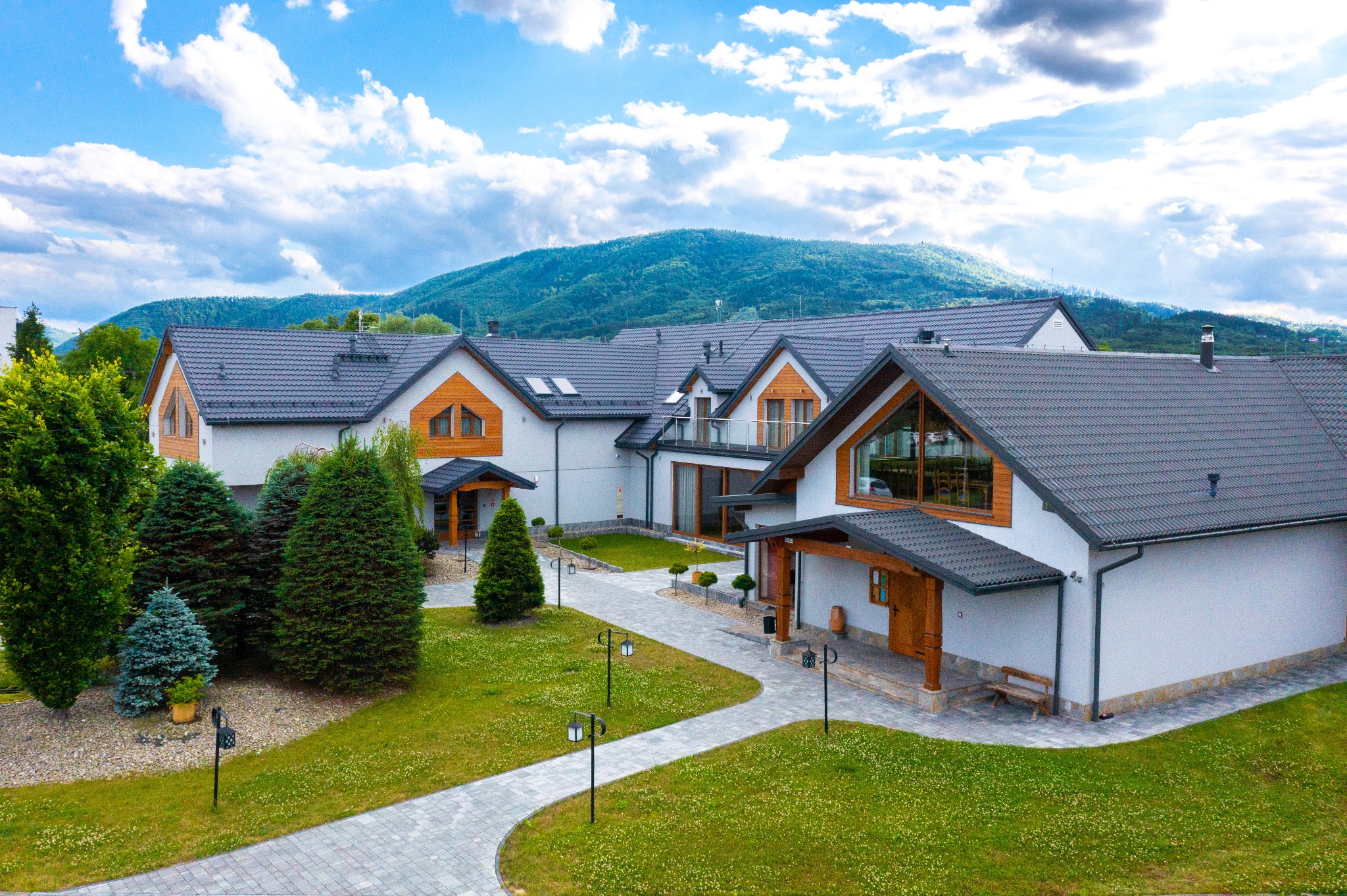 Rozległy, nowoczesny dom z szarą dachówką i jasną elewacją, otoczony zielonym trawnikiem, krzewami i drzewami iglastymi, z górą w tle pod błękitnym niebem z chmurami.