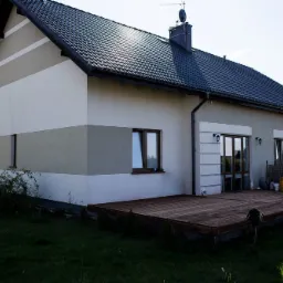 Widok z zewnątrz na dom jednorodzinny z ciemną dachówką, jasną elewacją w poziome pasy i drewnianym tarasem.