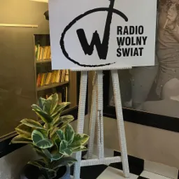 Logo 'Radio Wolny Świat' na białej planszy ustawionej na białej sztaludze, obok doniczka z zieloną rośliną, na podłodze czarno-biała szachownica.