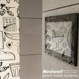 Panel sterowania Nexwell z wyświetlanym zegarem i graficznymi ikonami, zamontowany na szarej ścianie, obok ściany z białą tapetą w czarne rysunki.