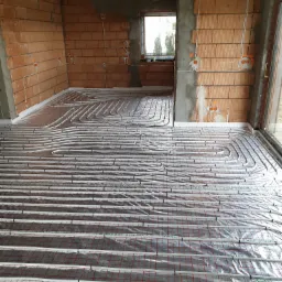 Rozłożone rury ogrzewania podłogowego na folii z siatką w surowym wnętrzu z ceglanymi ścianami i widocznymi instalacjami elektrycznymi.