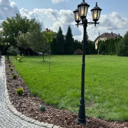 Oświetlony, czarny, trójramienny lampion ogrodowy na tle zielonego trawnika, rabaty z korą i kostki brukowej, z widocznymi drzewami i domem w tle.