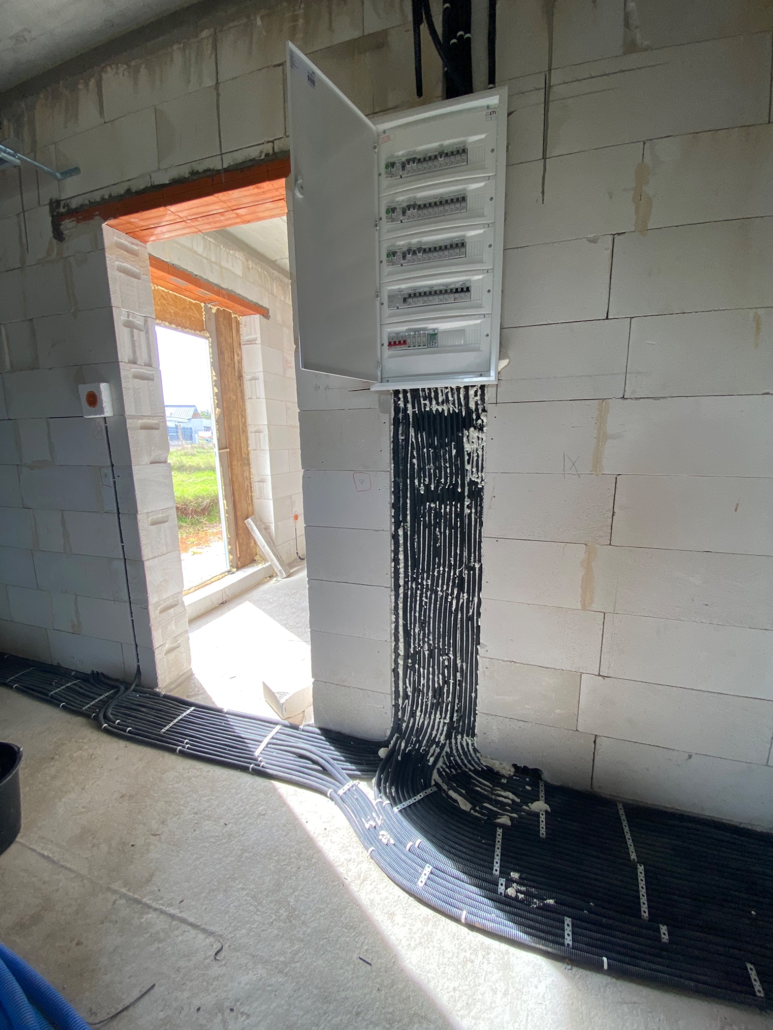 Rozdzielnica elektryczna z podłączonymi przewodami w peszlach, montowana na ścianie z białych bloczków w surowym stanie, widoczne mocowania przewodów do podłoża.