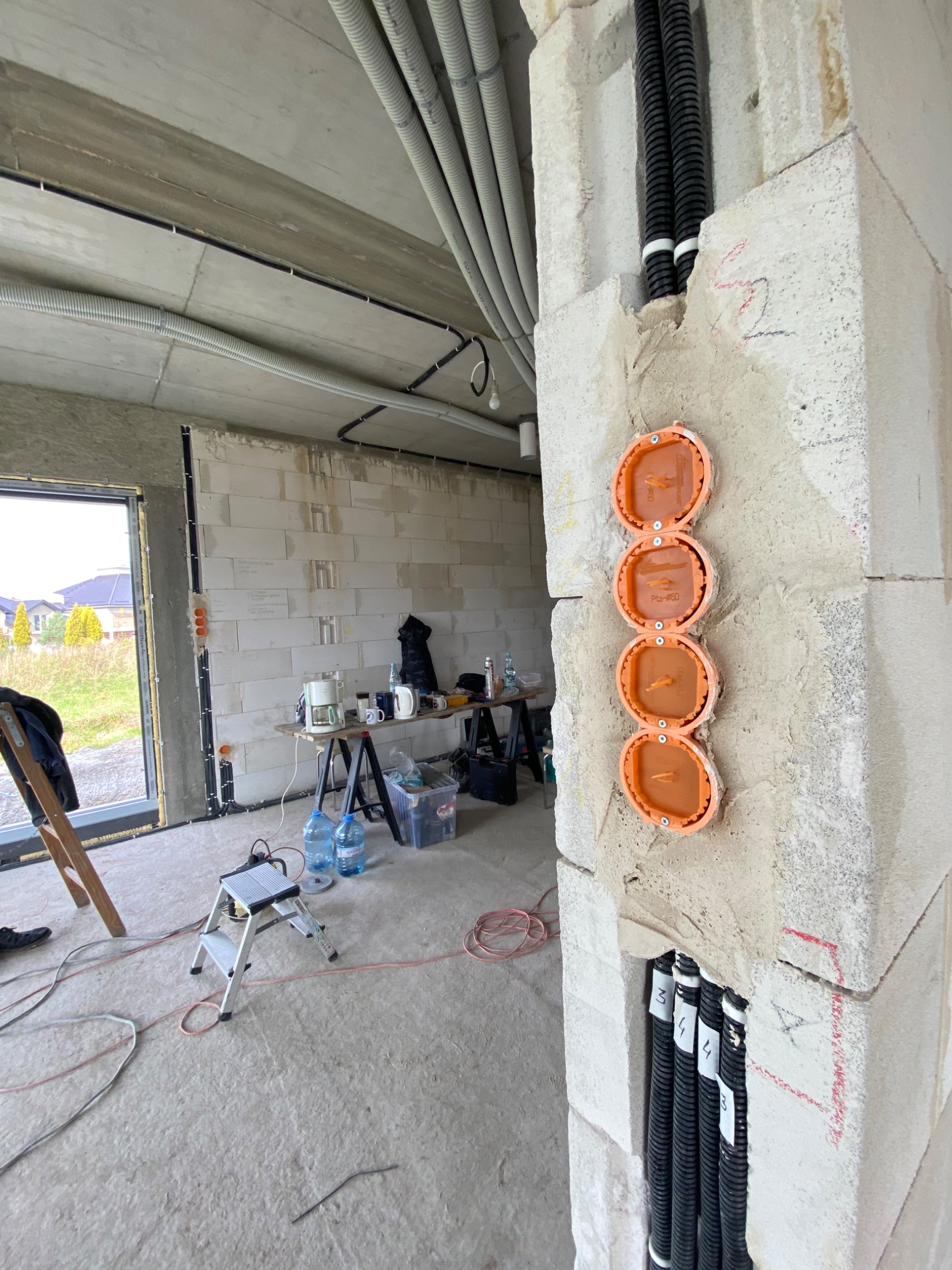 Wewnętrzna ściana z betonu komórkowego w trakcie prac elektrycznych: widoczne cztery pomarańczowe puszki instalacyjne w pionowym rzędzie, peszle z kablami, instalacja podtynkowa, surowy stan...