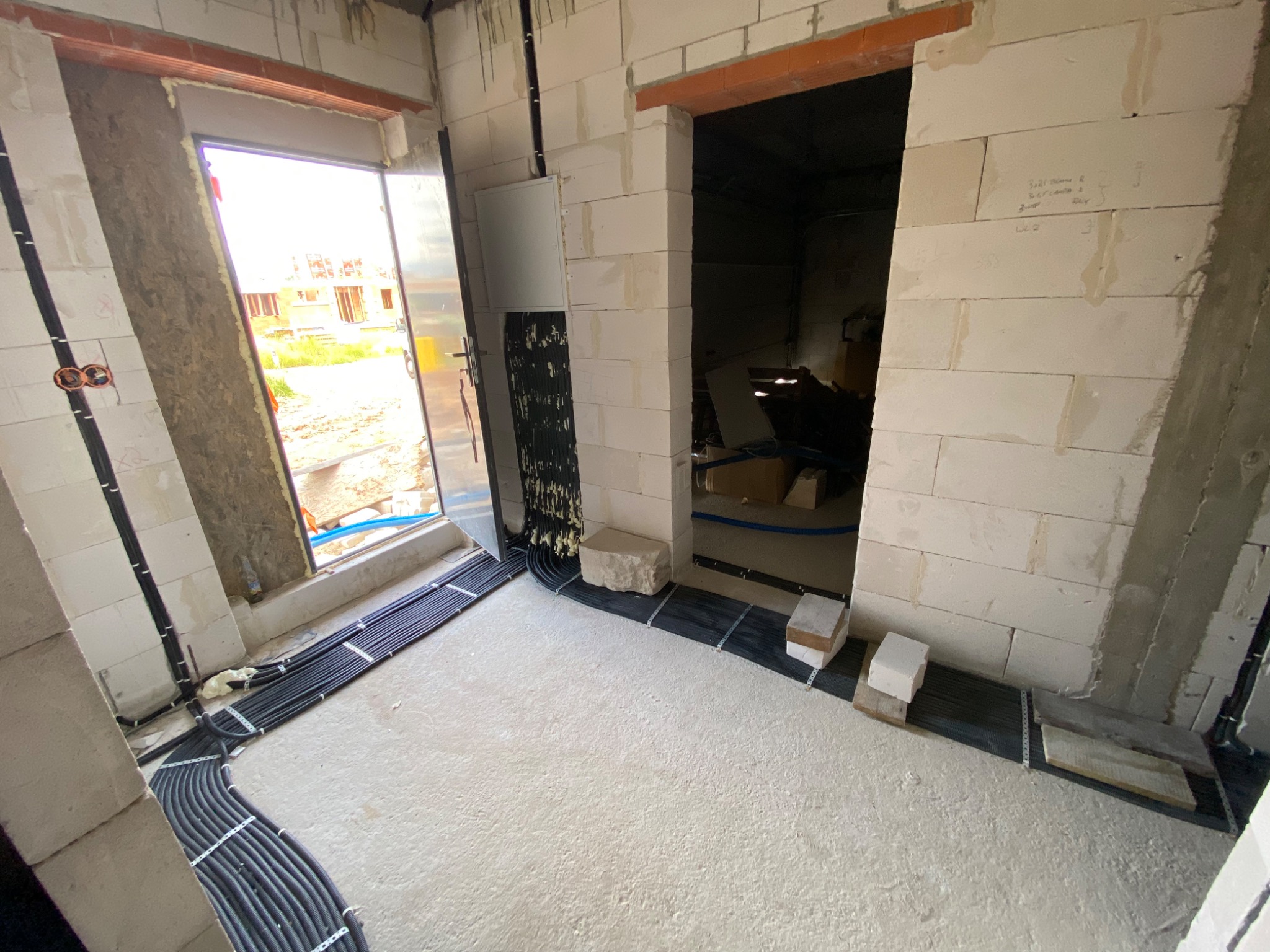 Wnętrze w stanie surowym: wiązki czarnych kabli elektrycznych ułożone na podłodze wzdłuż ścian, mocowane metalowymi uchwytami, widoczne ściany z białych bloczków i otwory drzwiowe.