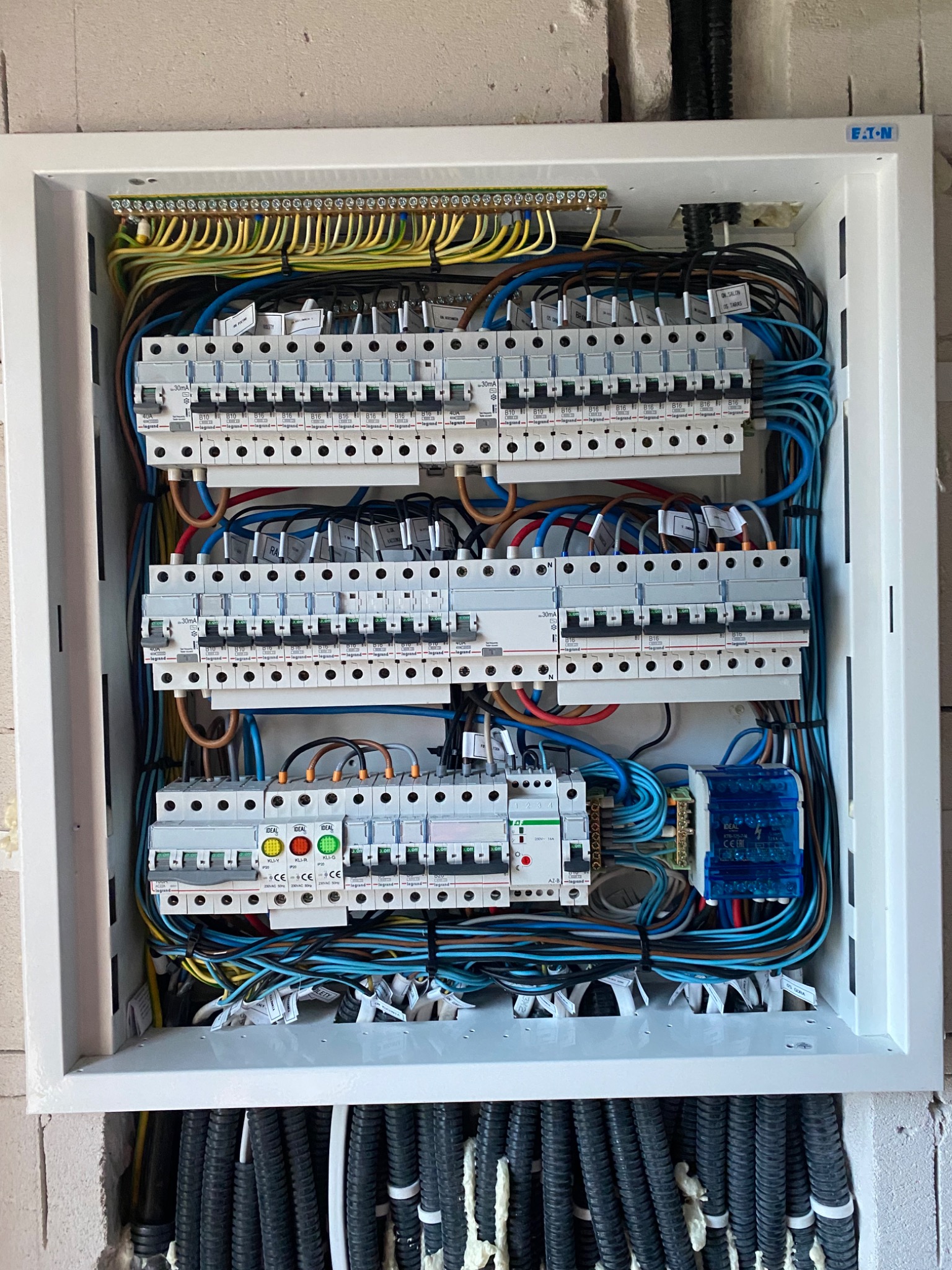 Otwarta skrzynka elektryczna z widocznymi bezpiecznikami, przewodami i złączkami, zamontowana na ścianie z widocznym logo firmy Eaton w prawym górnym rogu. Dolna część skrzynki wypełniona czarnymi,...