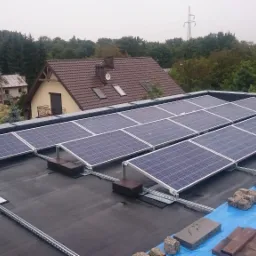 Montaż paneli fotowoltaicznych na płaskim dachu pokrytym papą, widoczne elementy konstrukcji wsporczej, cegły i deski zabezpieczające hydroizolację, w tle zabudowa miejska i drzewa.