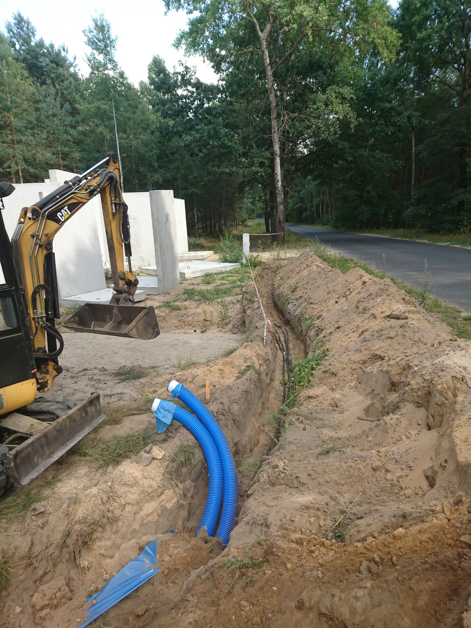 Wykop pod ziemią z ułożonymi niebieskimi rurami karbowanymi, obok koparka CAT i betonowe fundamenty, w tle las i asfaltowa droga.