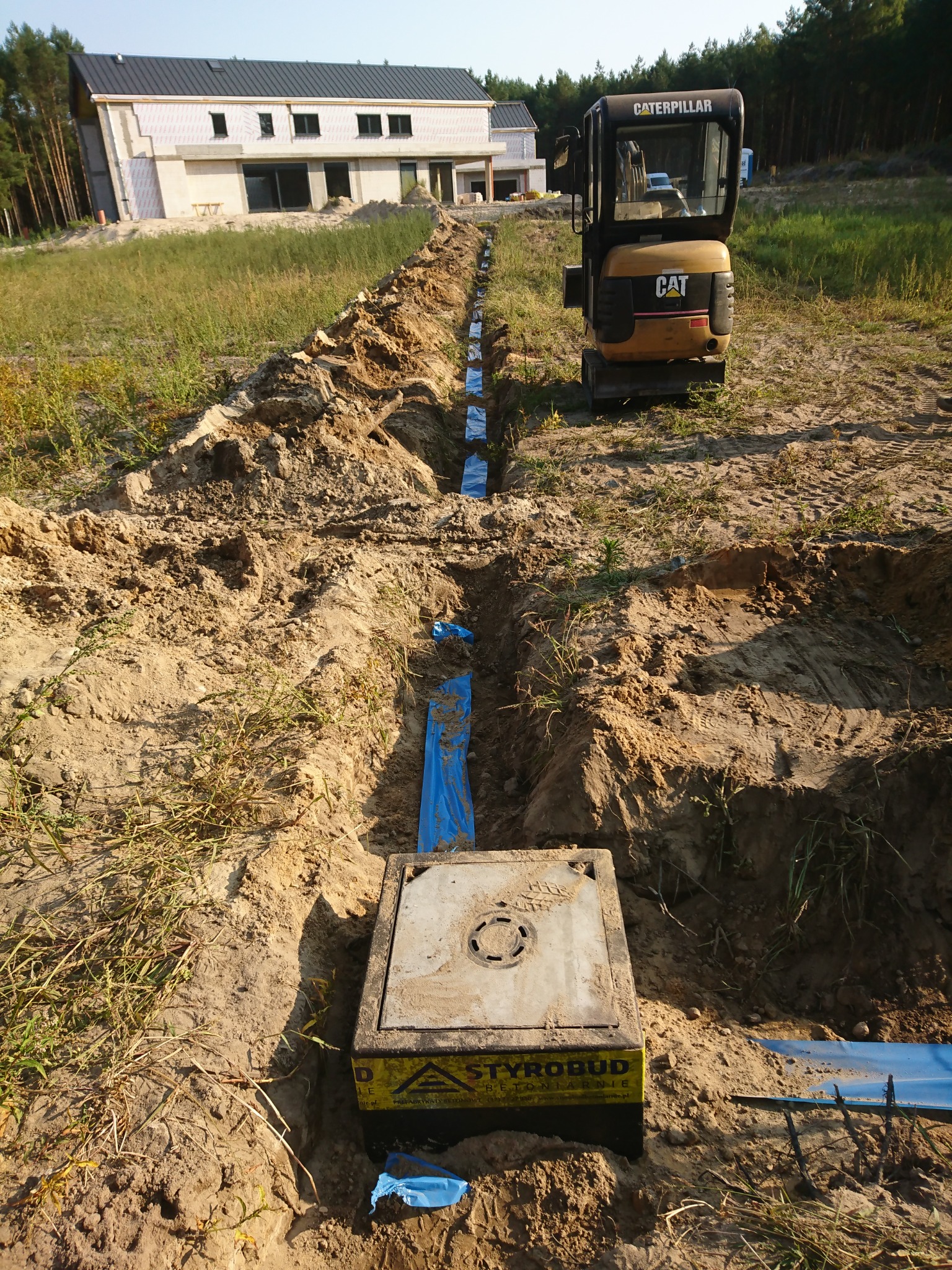 Wykopany rów pod przyłącze elektryczne z widoczną studzienką kablową Styrobud, niebieską folią ochronną i minikoparką Caterpillar w tle, na tle domu w budowie i lasu.