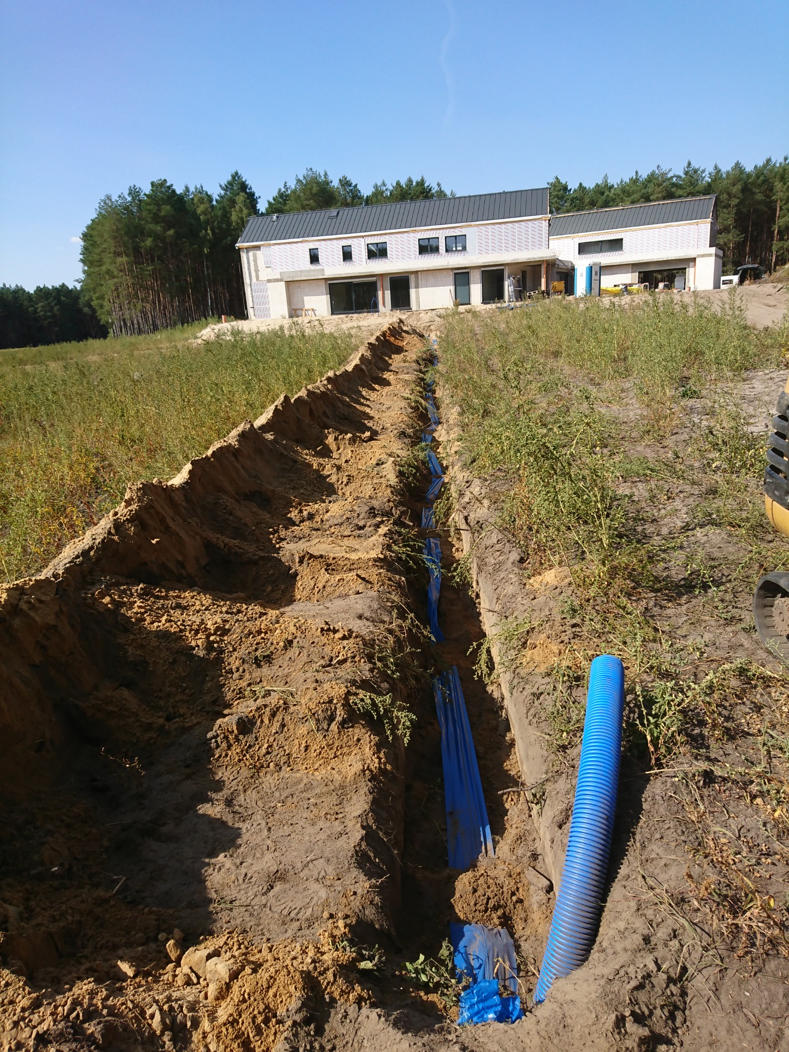 Wykop w ziemi z ułożonymi niebieskimi rurami karbowanymi, widok na dom w budowie i las w tle, słoneczny dzień.