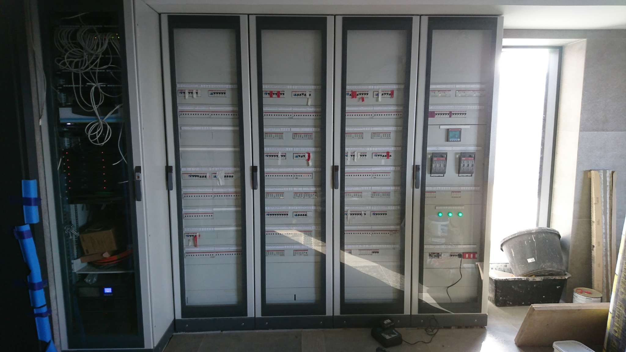 Rozdzielnica elektryczna z czterema segmentami, z otwartą szafą rackową obok, widoczne kable i elementy montażowe, na podłodze narzędzia i akcesoria budowlane, światło dzienne z okna.