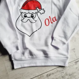 Biała bluza z nadrukiem Świętego Mikołaja i imieniem 'Ola' w kolorze czerwonym, leżąca na drewnianym tle.