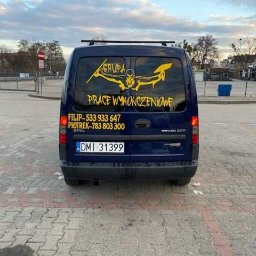 Rad Mir Godek Radosław - Tył granatowego Opla Combo z żółtym nadrukiem 'GRUPA PRACE WYKOŃCZENIOWE' oraz numerami telefonów na tylnej szybie, zaparkowanego na brukowanym placu.