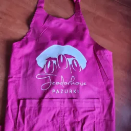 Różowy fartuch z białym nadrukiem logo 'Teodorowe Pazurki' leżący na drewnianym stole.