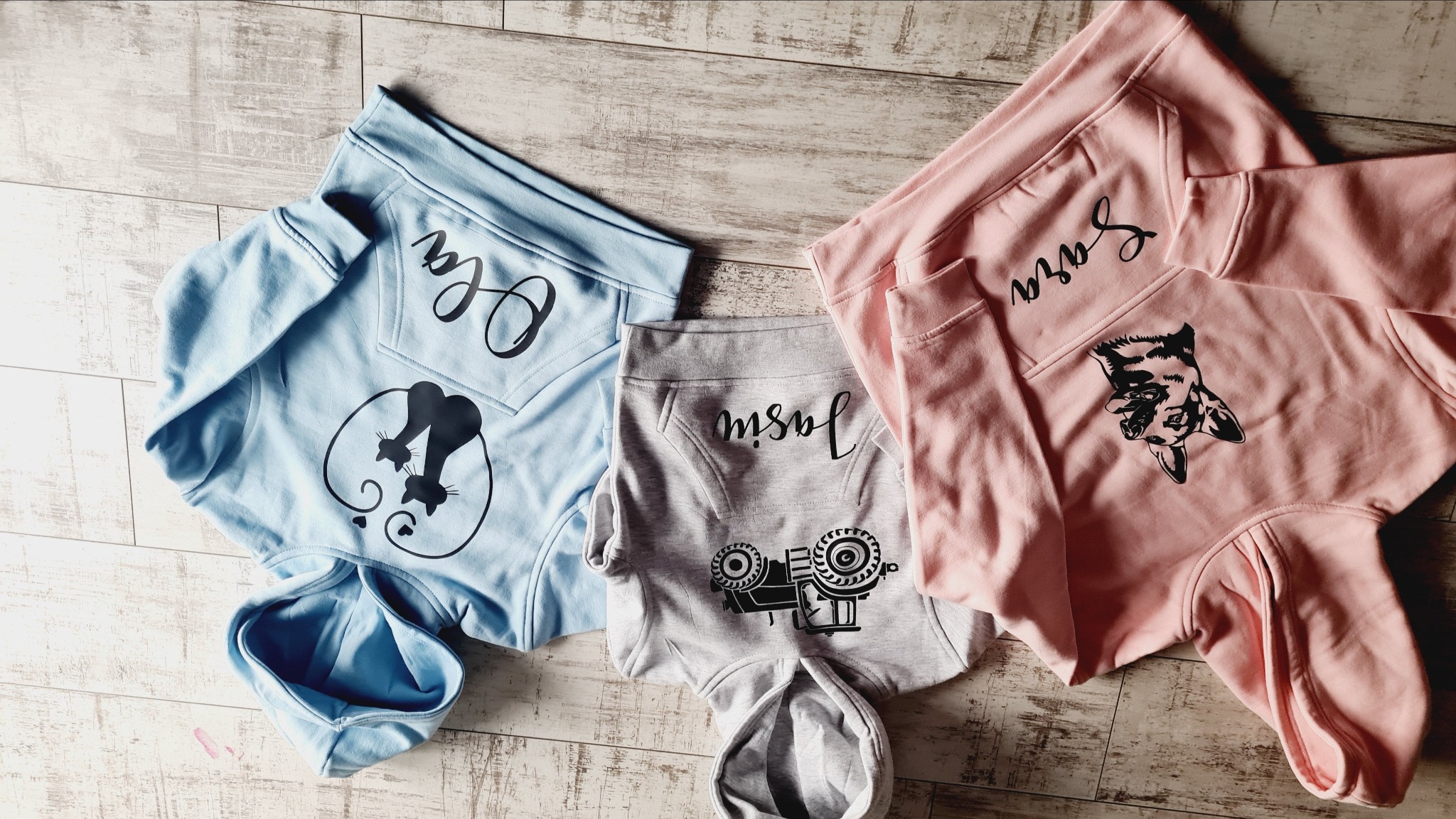 Trzy bluzy z kapturem w kolorach pastelowych (różowy, błękitny, szary) leżące na drewnianym podłożu; każda z bluz ma nadruk z imieniem i grafiką (koty, pies, traktor).