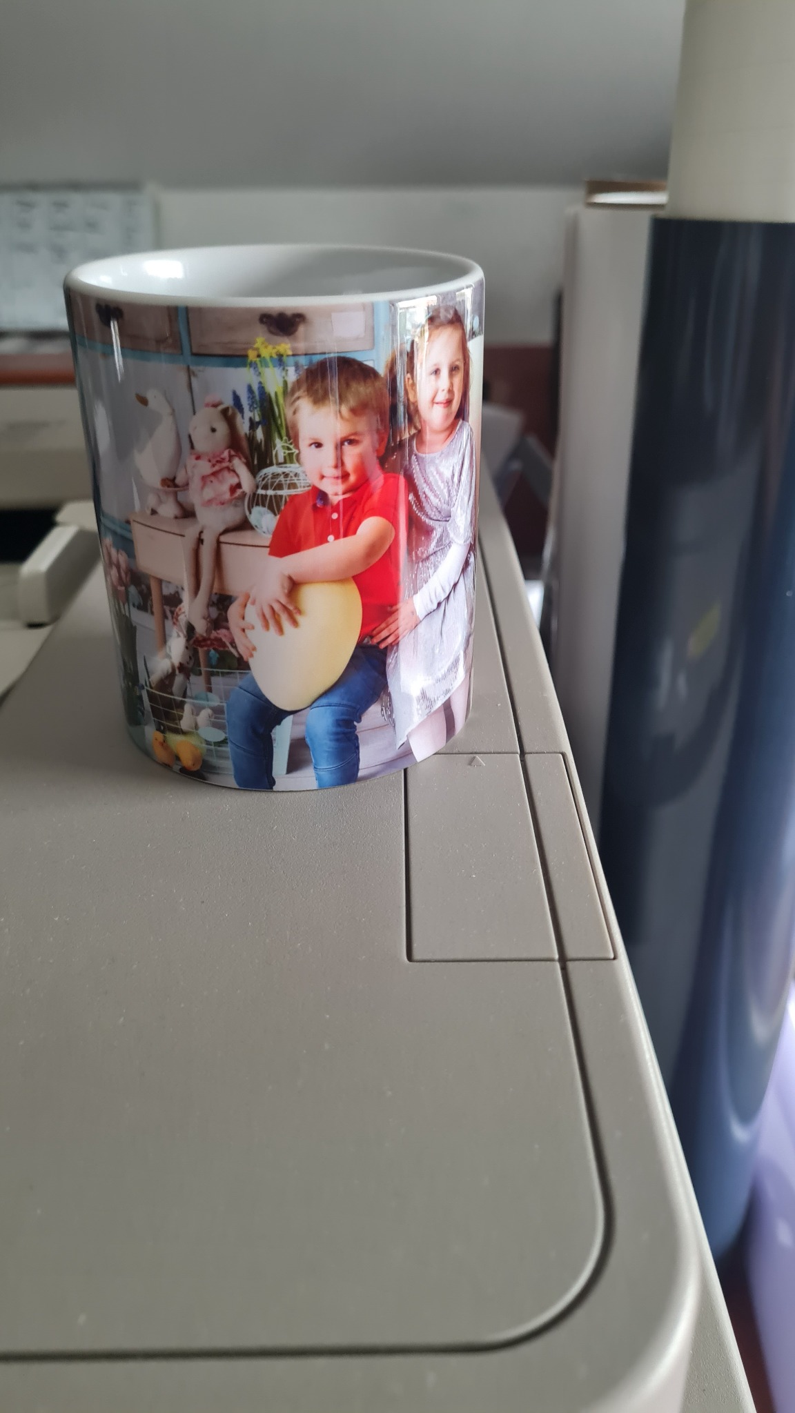 Kubek z nadrukiem zdjęcia dwójki dzieci, stojący na szarym urządzeniu drukującym.