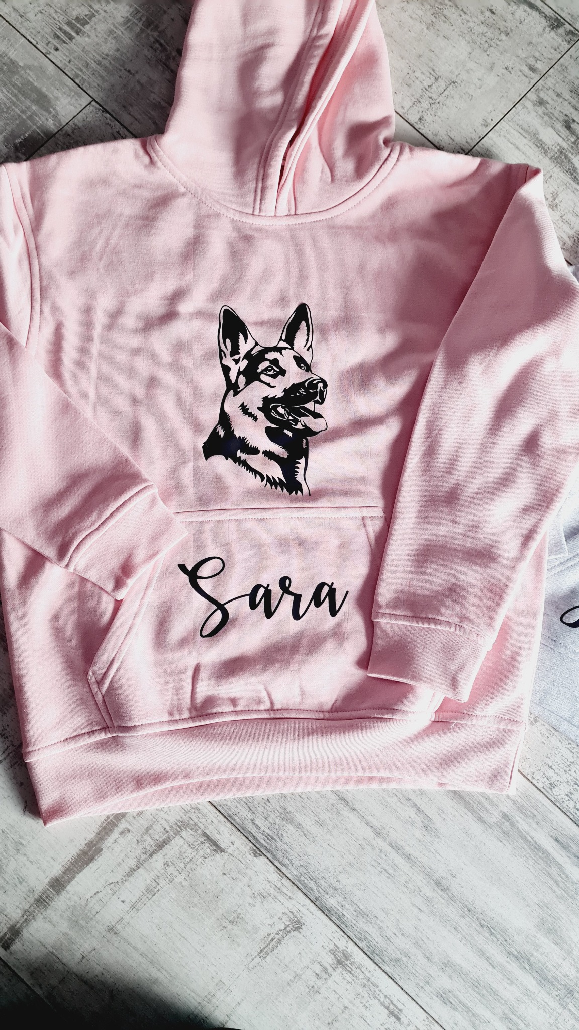 Różowa bluza z kapturem z czarnym nadrukiem głowy owczarka niemieckiego i imieniem 'Sara' na kieszeni, leżąca na drewnianym tle.