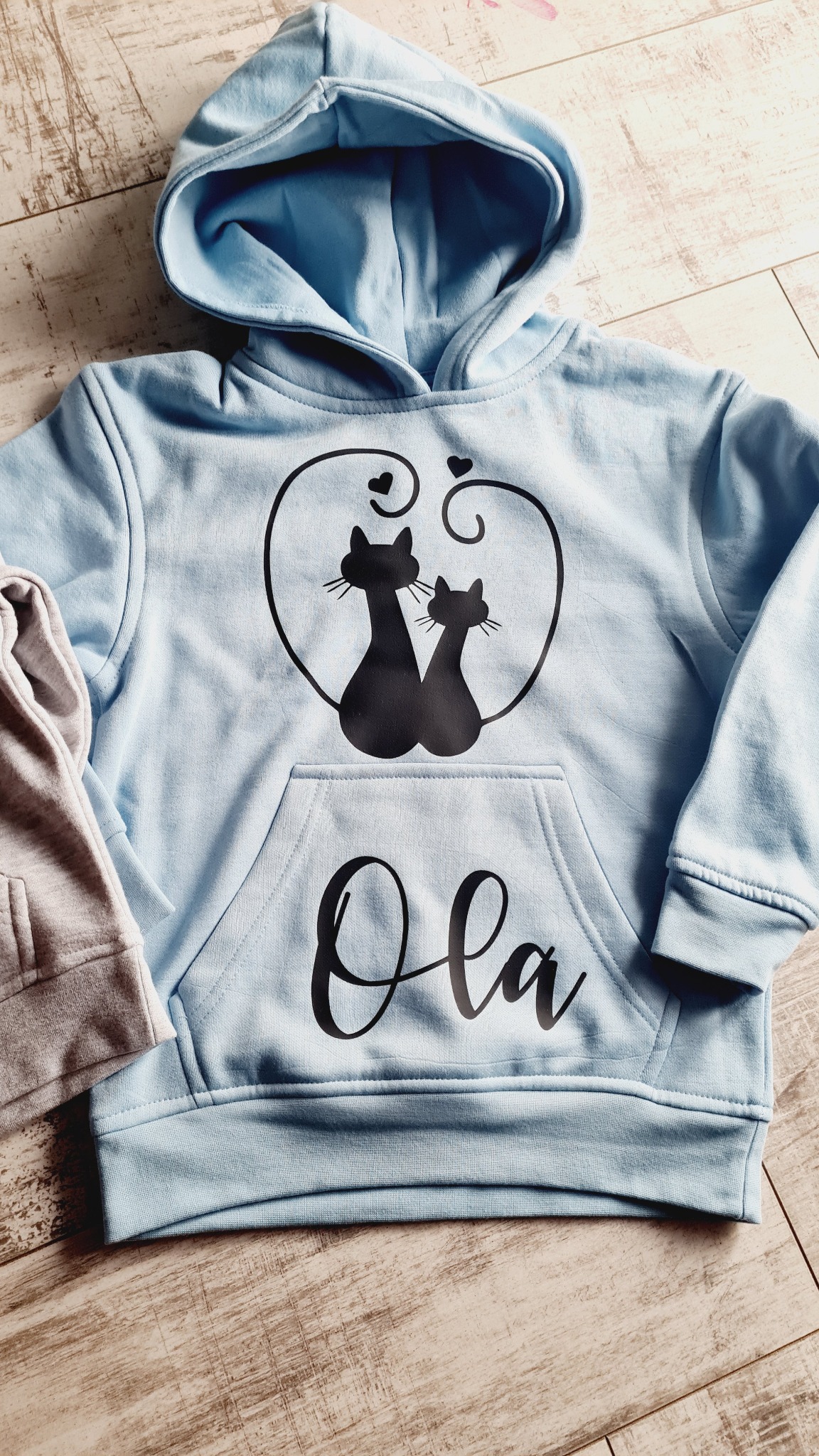 Jasnoniebieska bluza z kapturem, z nadrukiem dwóch czarnych kotów w sercu i imieniem 'Ola' na kieszeni, leżąca na drewnianym tle obok szarej bluzy.