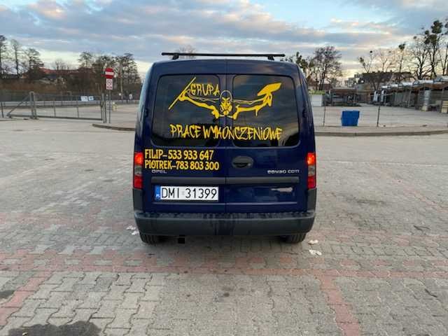 Tył granatowego Opla Combo z żółtym nadrukiem 'GRUPA PRACE WYKOŃCZENIOWE' oraz numerami telefonów na tylnej szybie, zaparkowanego na brukowanym placu.