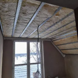 Poddasze w trakcie ocieplania, widoczne wełna mineralna między metalowymi profilami sufitowymi, okno z widokiem na zimowy krajobraz.