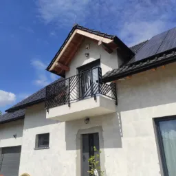 Nowoczesny dom z balkonem z czarną balustradą o geometrycznym wzorze, widok z dołu na tle błękitnego nieba z lekkimi chmurami.