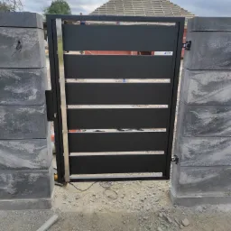 Nowoczesna brama wjazdowa z poziomymi panelami w kolorze czarnym, osadzona na szarych, betonowych słupkach, widok na plac budowy i fragment dachu domu.