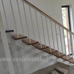 Schody metalowe z drewnianymi stopniami i białą balustradą w trakcie montażu, zabezpieczone niebieską taśmą malarską, widoczne elementy wykończeniowe podłogi.