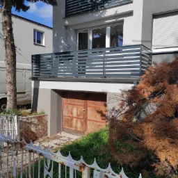 Szara balustrada balkonowa z poziomych profili, widok z zewnątrz, zardzewiałe drzwi garażowe poniżej, białe ogrodzenie metalowe w tle, częściowo zasłonięte przez brązowy krzew.