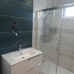 Nowoczesna łazienka z ciemnozielonymi płytkami na ścianie, białą szafką pod umywalką z trzema szufladami i kabiną prysznicową z przezroczystego szkła, widoczne niedokończone podłączenie elektryczne...