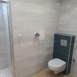 Wykończona łazienka z szarymi płytkami imitującymi kamień, kabiną prysznicową ze srebrnymi profilami i toaletą podwieszaną z zabudową w kolorze ciemnego turkusu.