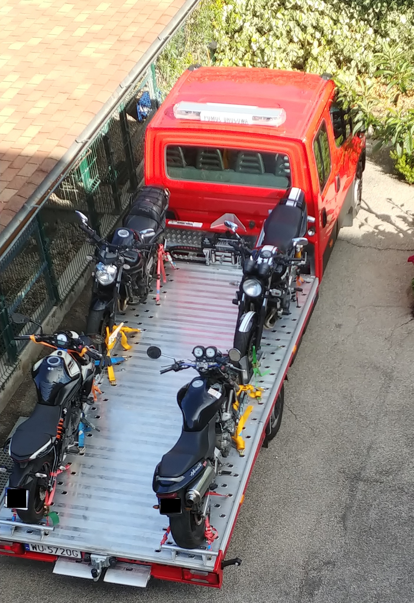 Czerwona laweta z czterema motocyklami zabezpieczonymi pasami transportowymi, widok z góry na tle zieleni i dachu budynku.