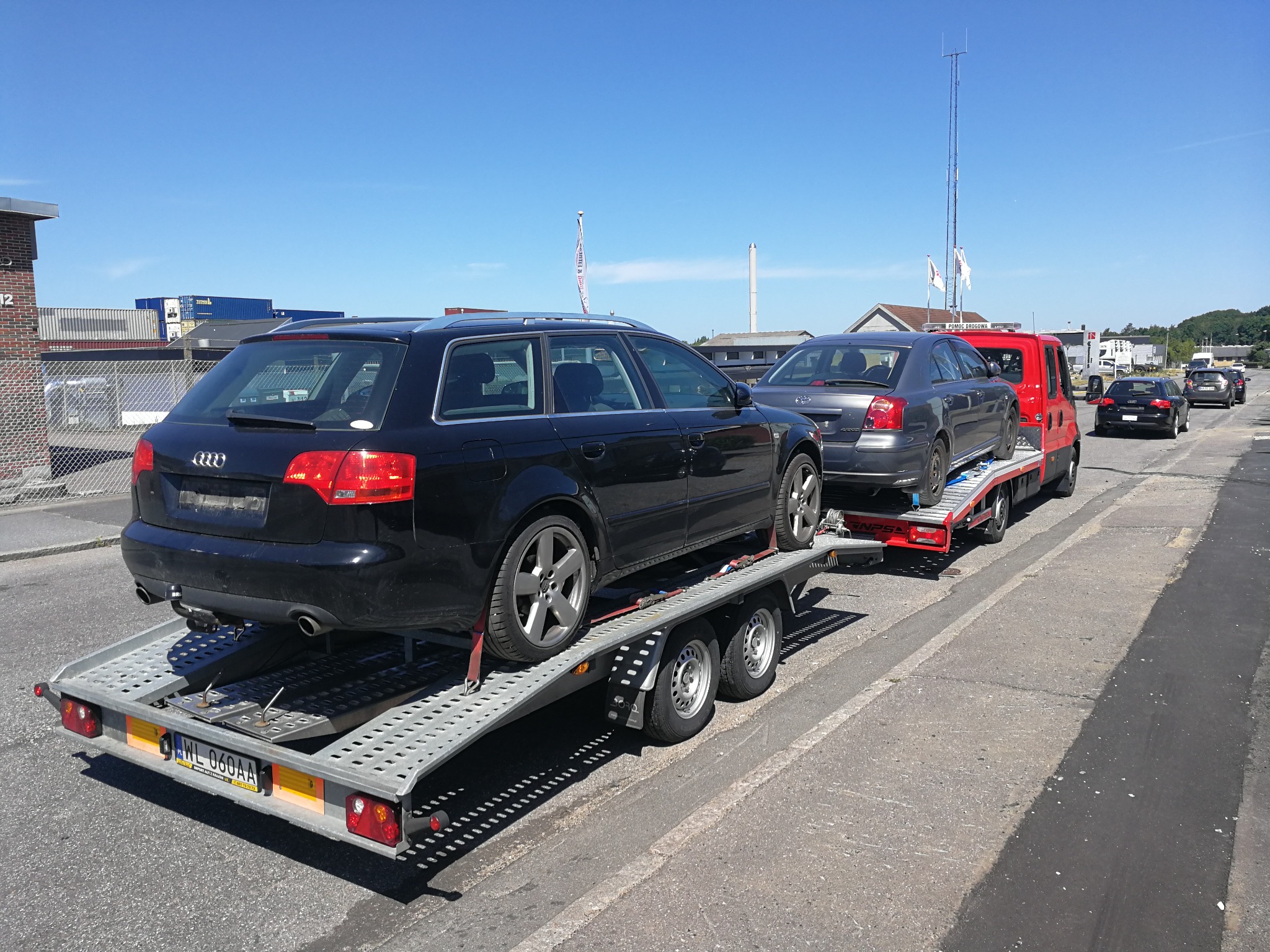 Czarny samochód Audi A4 Avant na przyczepie lawetowej, widok z tyłu, tablica rejestracyjna WL 060AA, w tle czerwona laweta z drugim samochodem i słup telekomunikacyjny