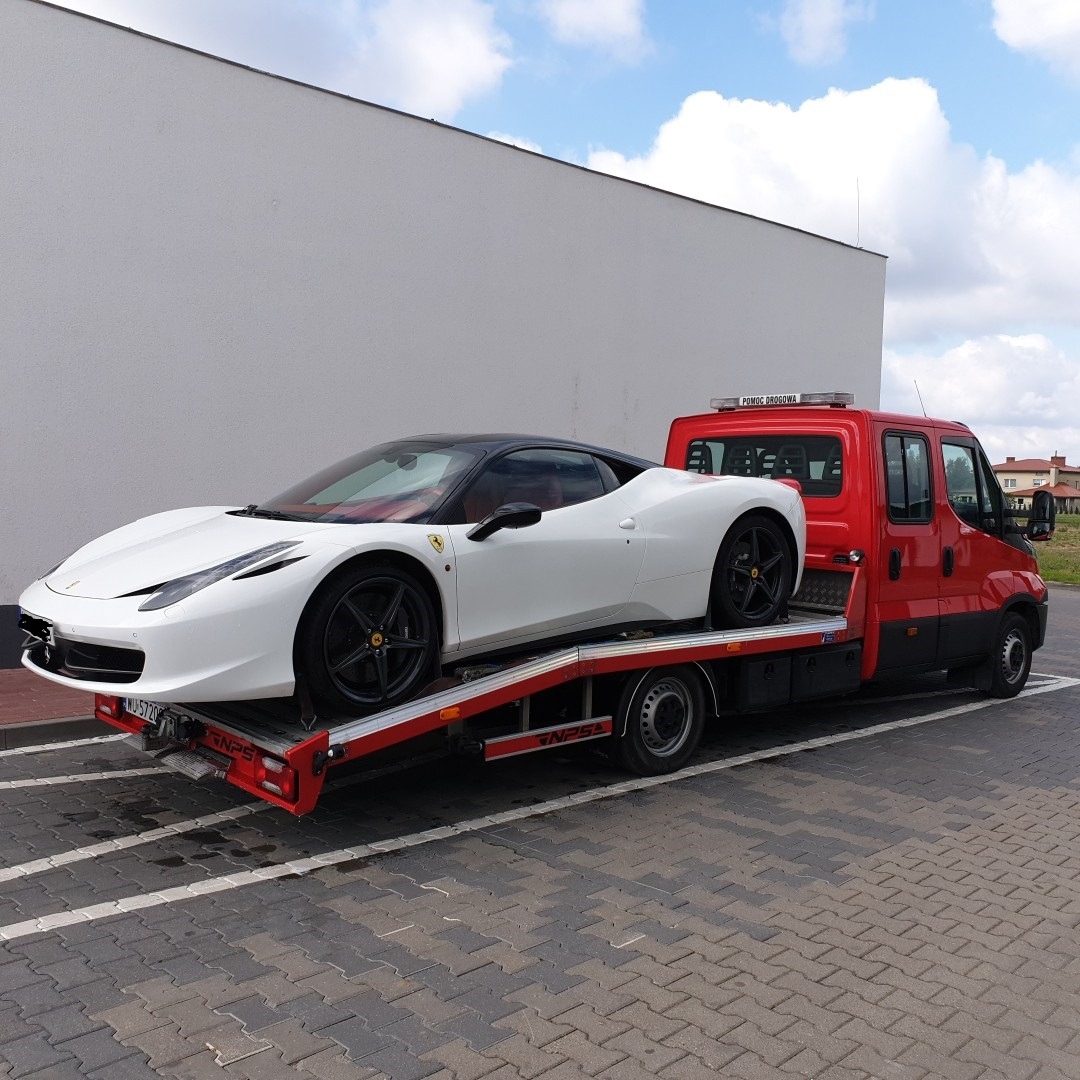 Białe Ferrari 458 Italia na czerwonej lawecie pomocy drogowej Iveco, widok z boku na parkingu z kostki brukowej.