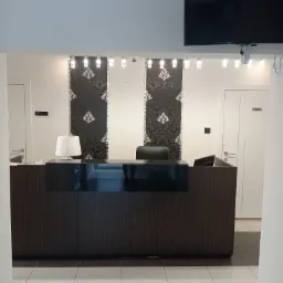 Recepcja z ciemnego drewna z czarnym blatem, oświetlona lampą stołową, z dekoracyjnymi panelami ściennymi w tle, oświetleniem sufitowym i telewizorem na ścianie.