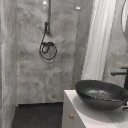 Nowoczesna łazienka z szarą, betonową płytką, czarną armaturą prysznicową i umywalką nablatową w kształcie misy, biała szafka pod umywalką z minimalistycznymi uchwytami.