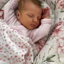 Śpiące niemowlę w różowym ubranku, przykryte białą pieluszką w różowe kwiatki, leżące w kokonie z motywem piwonii.