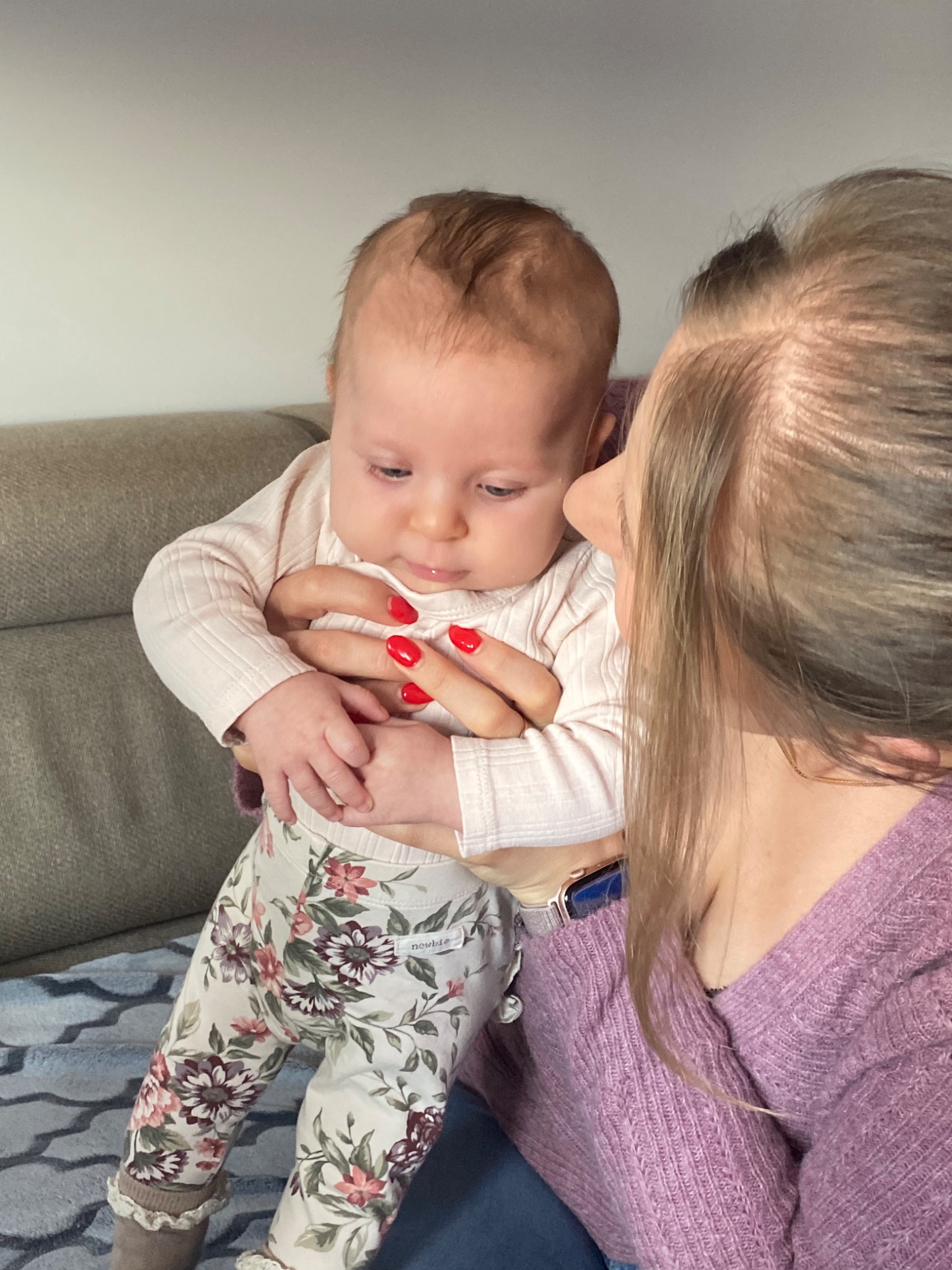 Kobieta z czerwonymi paznokciami trzyma niemowlę w kwiecistych legginsach na tle szarej kanapy.