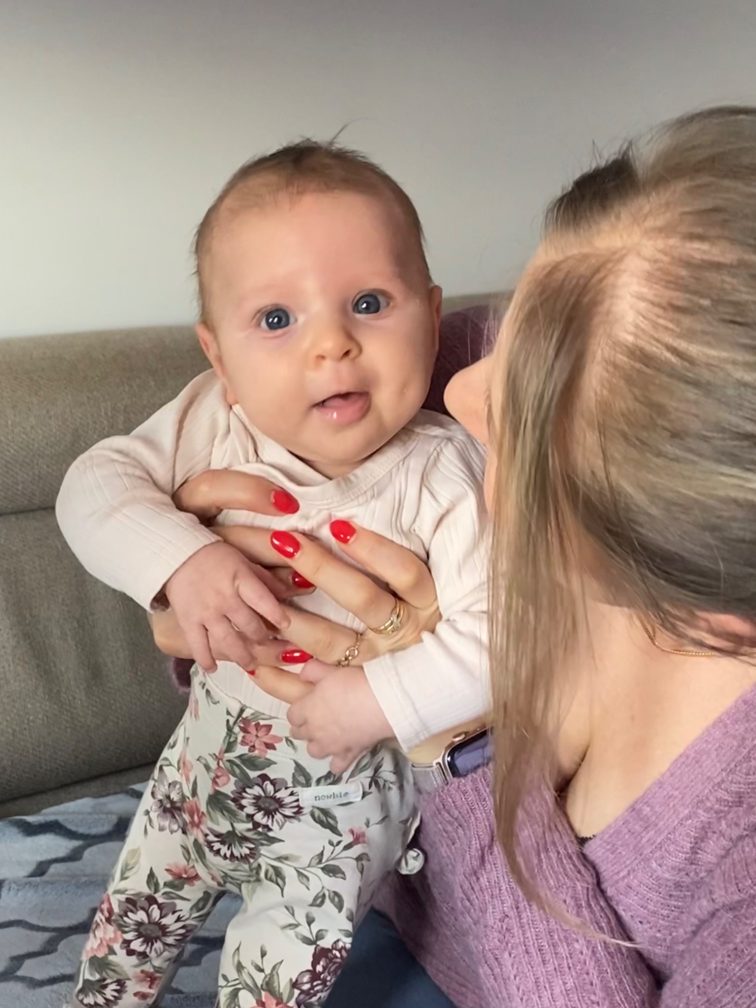 Uśmiechnięte niemowlę w kwiatowych legginsach trzymane przez kobietę z czerwonymi paznokciami na tle beżowej sofy.