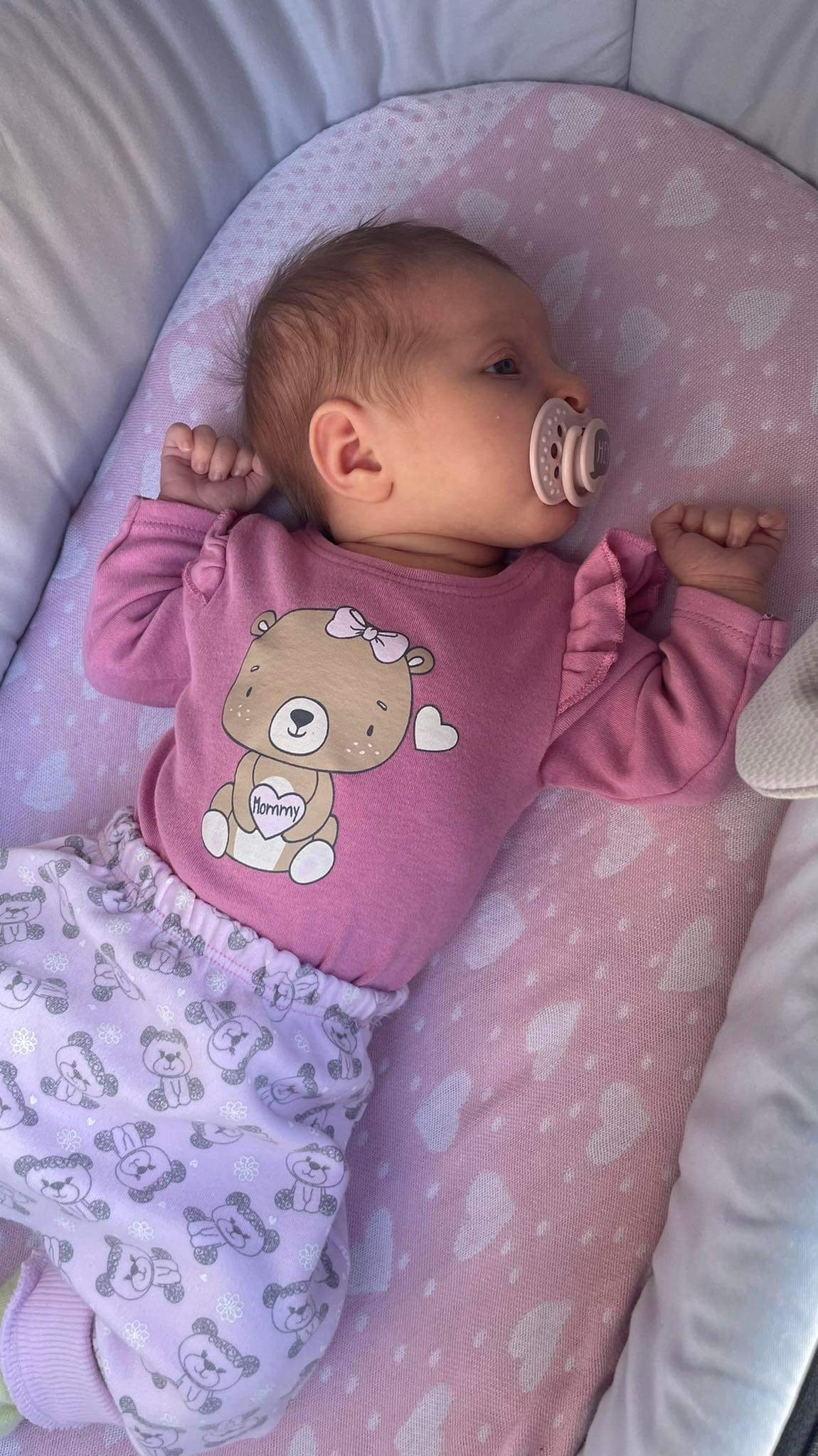 Niemowlę z smoczkiem leży na różowym kocyku w łóżeczku, ubrane w różowe ubranko z nadrukiem misia.