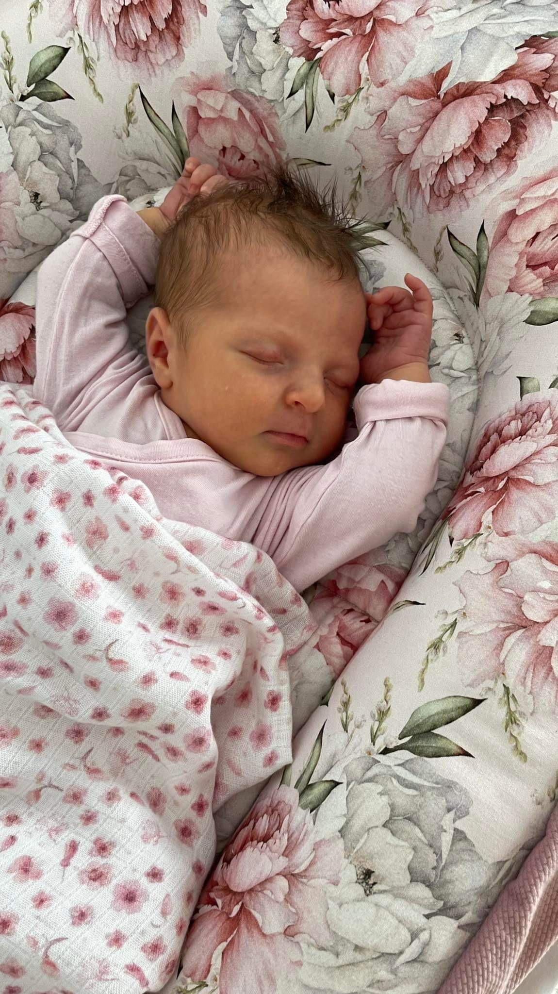 Śpiące niemowlę w różowym ubranku, przykryte białą pieluszką w różowe kwiatki, leżące w kokonie z motywem piwonii.