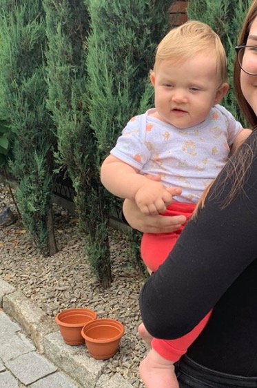 Kobieta trzyma na rękach małe dziecko w ogrodzie, obok stoją dwie puste doniczki.