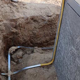 Elektros - Wykop z ułożonym metalowym uziomem pionowym i poziomym, zabezpieczonym żółtą osłoną. Widoczna ściana budynku i narzędzia w tle. Prace ziemne.