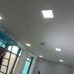 Elektros - Biały sufit z zamontowanymi, kwadratowymi lampami LED. Widoczne okna zabezpieczone folią i drabina. Ujęcie z dołu, podkreślające nowoczesne oświetlenie.