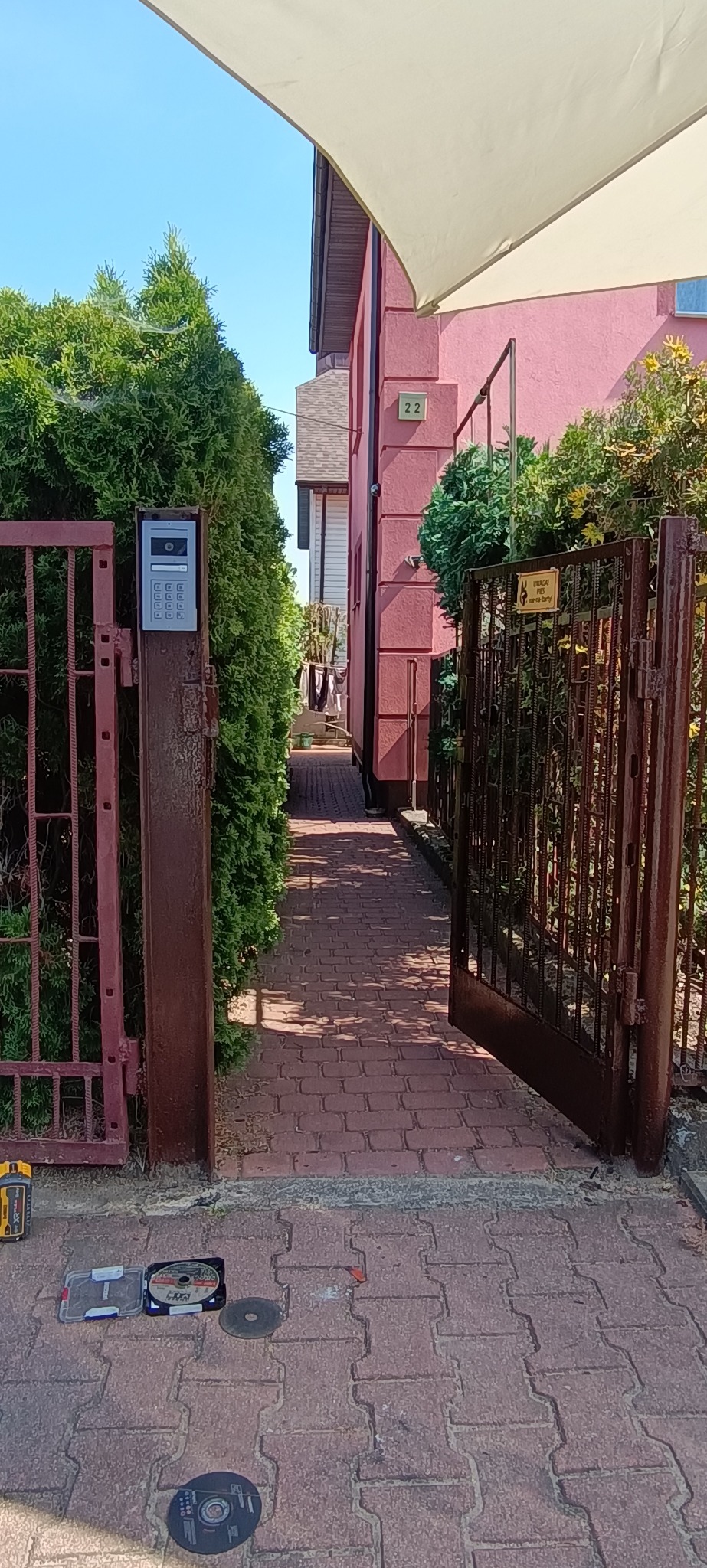 Otwarta, brązowa brama z domofonem przy posesji z różową elewacją. Na brukowanym podłożu widoczne narzędzia.