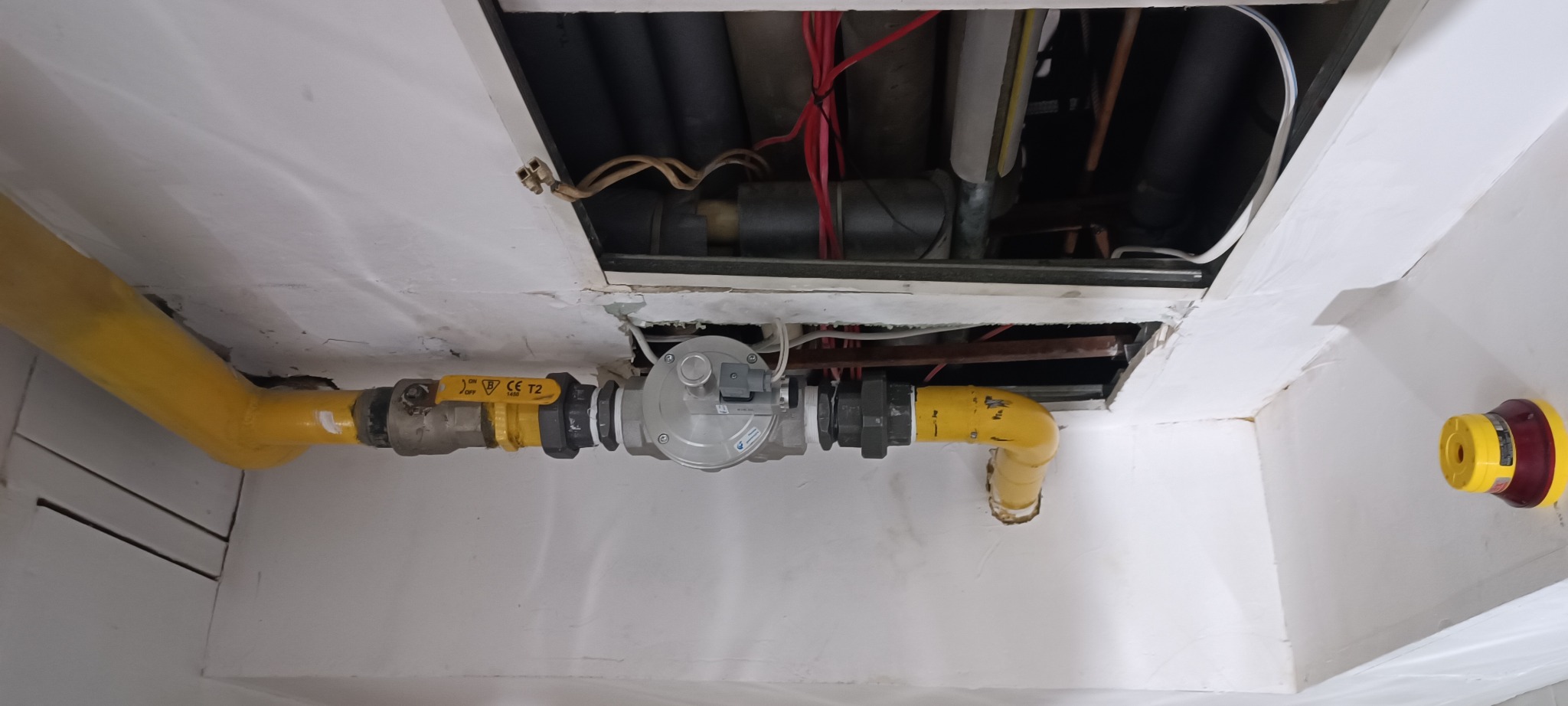 Żółta instalacja gazowa z regulatorem ciśnienia pod sufitem z widocznymi przewodami w otworze. Fragment ściany z białymi panelami.