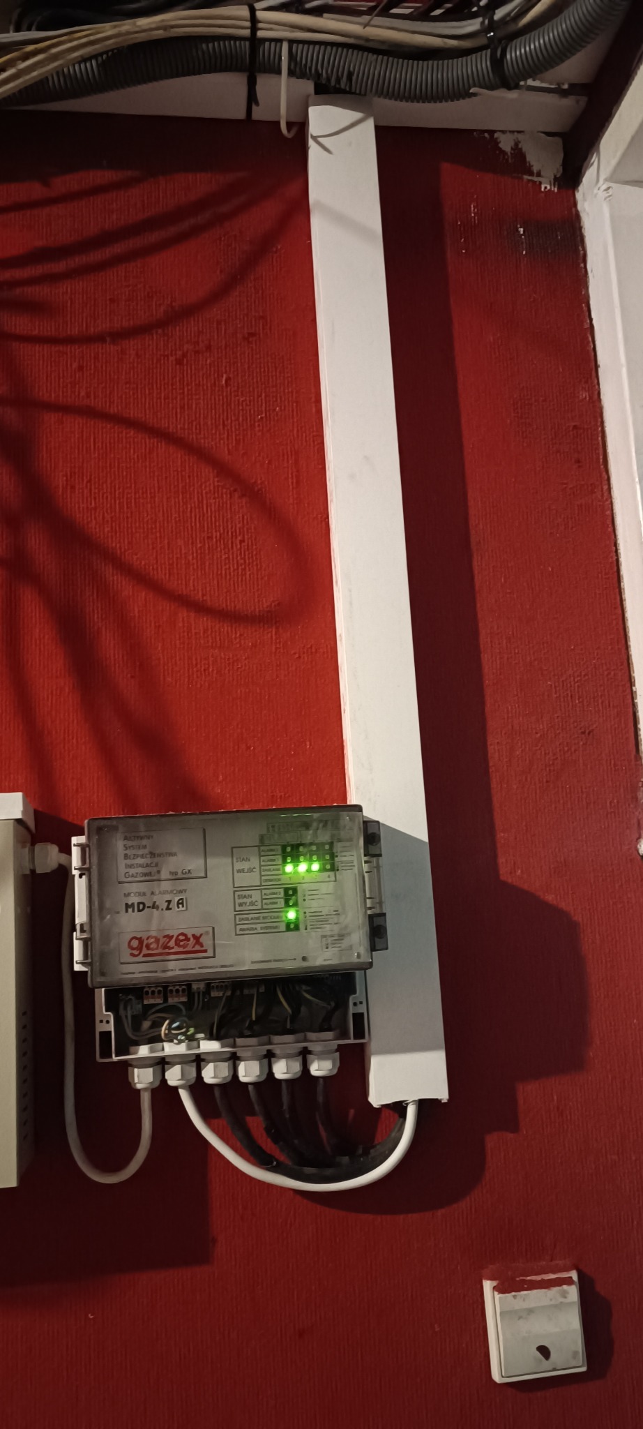 Alarmowy moduł Gazex MD-4.ZA na czerwonej ścianie, z widocznymi przewodami i diodami sygnalizacyjnymi. Element instalacji gazowej w budynku.