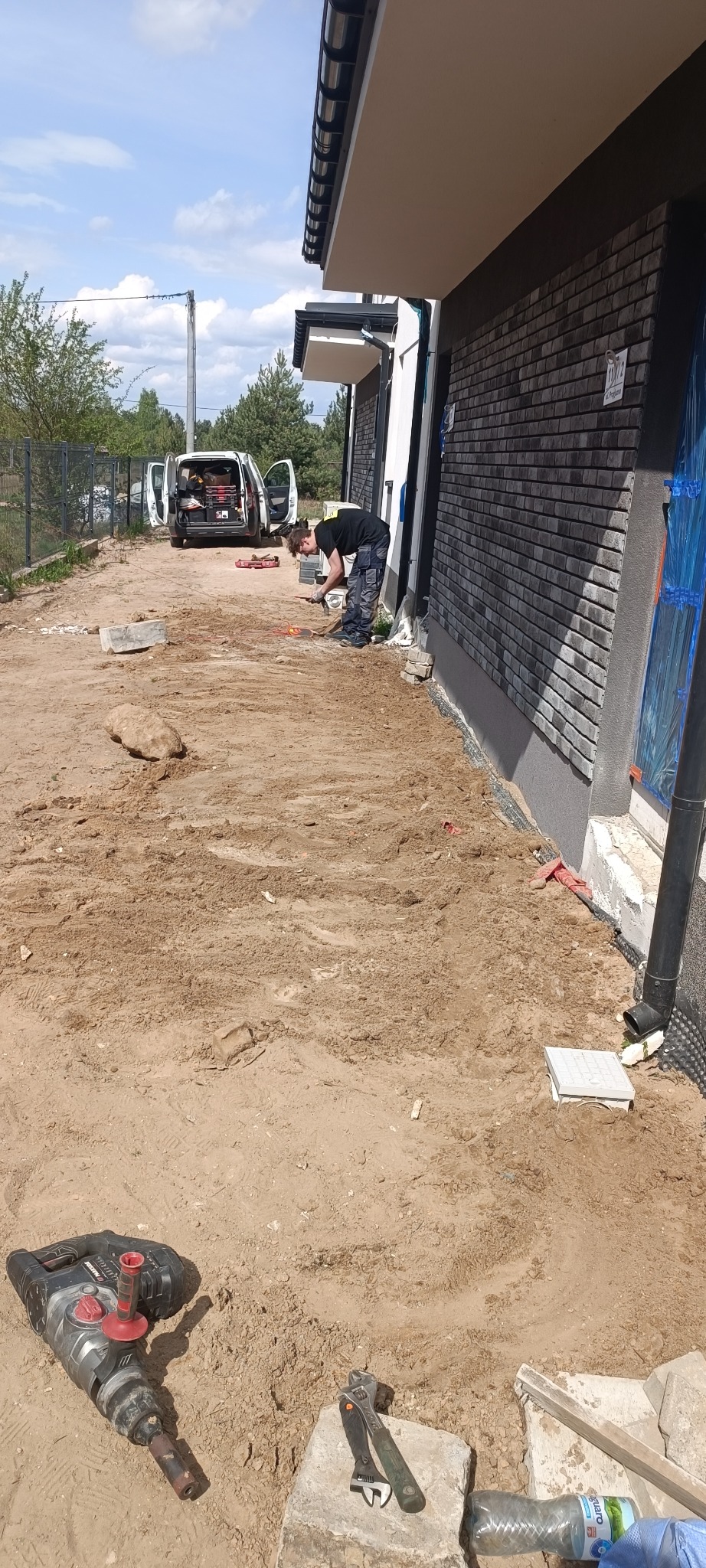 Pracownik w trakcie prac ziemnych przy budynku z cegły klinkierowej. Widoczne narzędzia, w tle biały samochód dostawczy z otwartymi drzwiami. Dzień słoneczny.