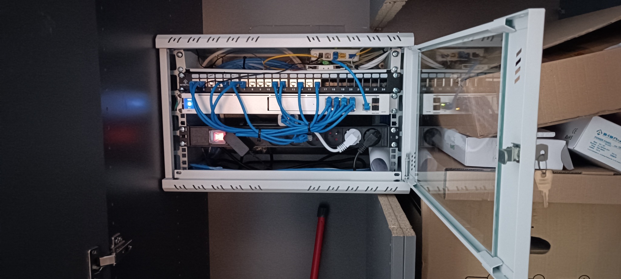 Otwarta szafka rack z zainstalowanym panelem krosowniczym, routerem i listwą zasilającą. Widoczne niebieskie kable sieciowe. Szafka zamontowana w ciemnej obudowie.