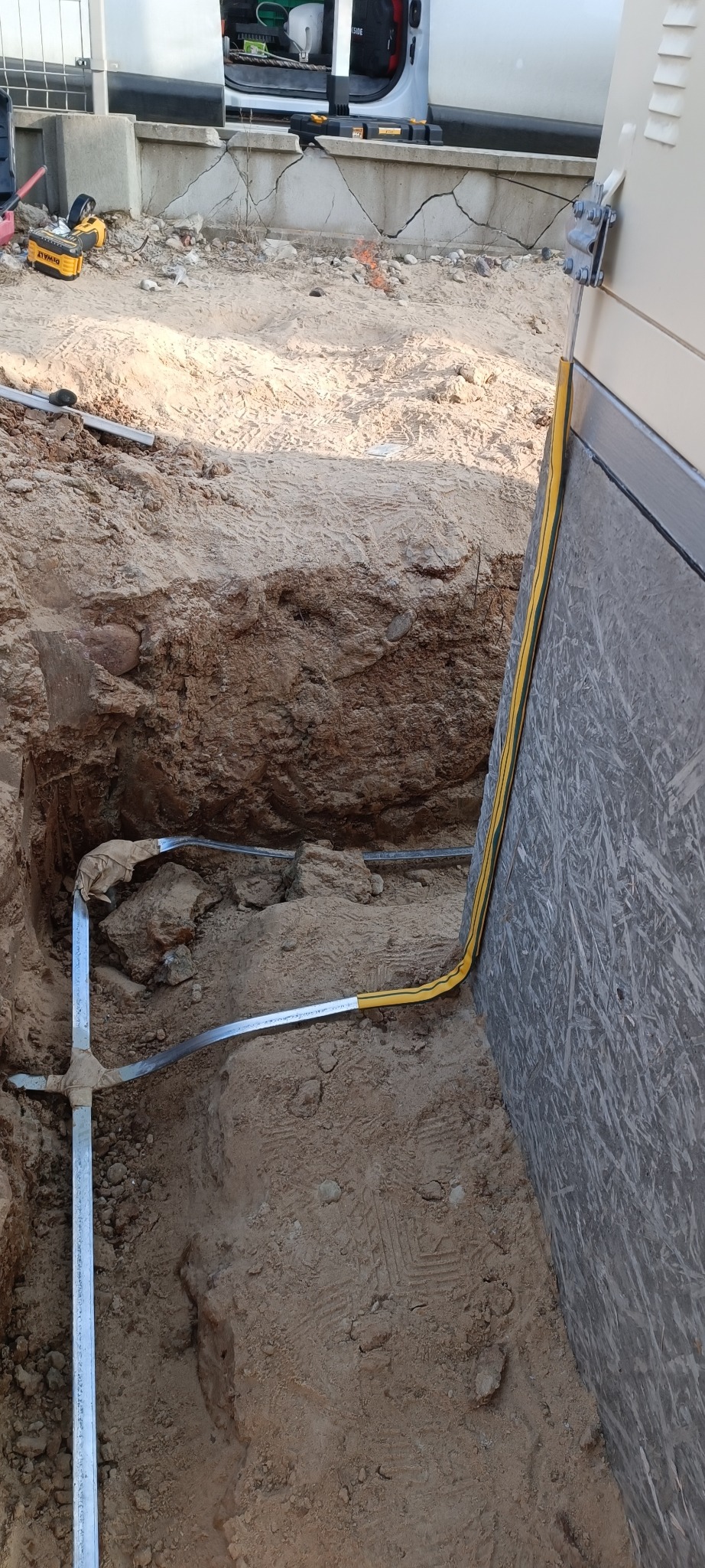 Wykop z ułożonym metalowym uziomem pionowym i poziomym, zabezpieczonym żółtą osłoną. Widoczna ściana budynku i narzędzia w tle. Prace ziemne.