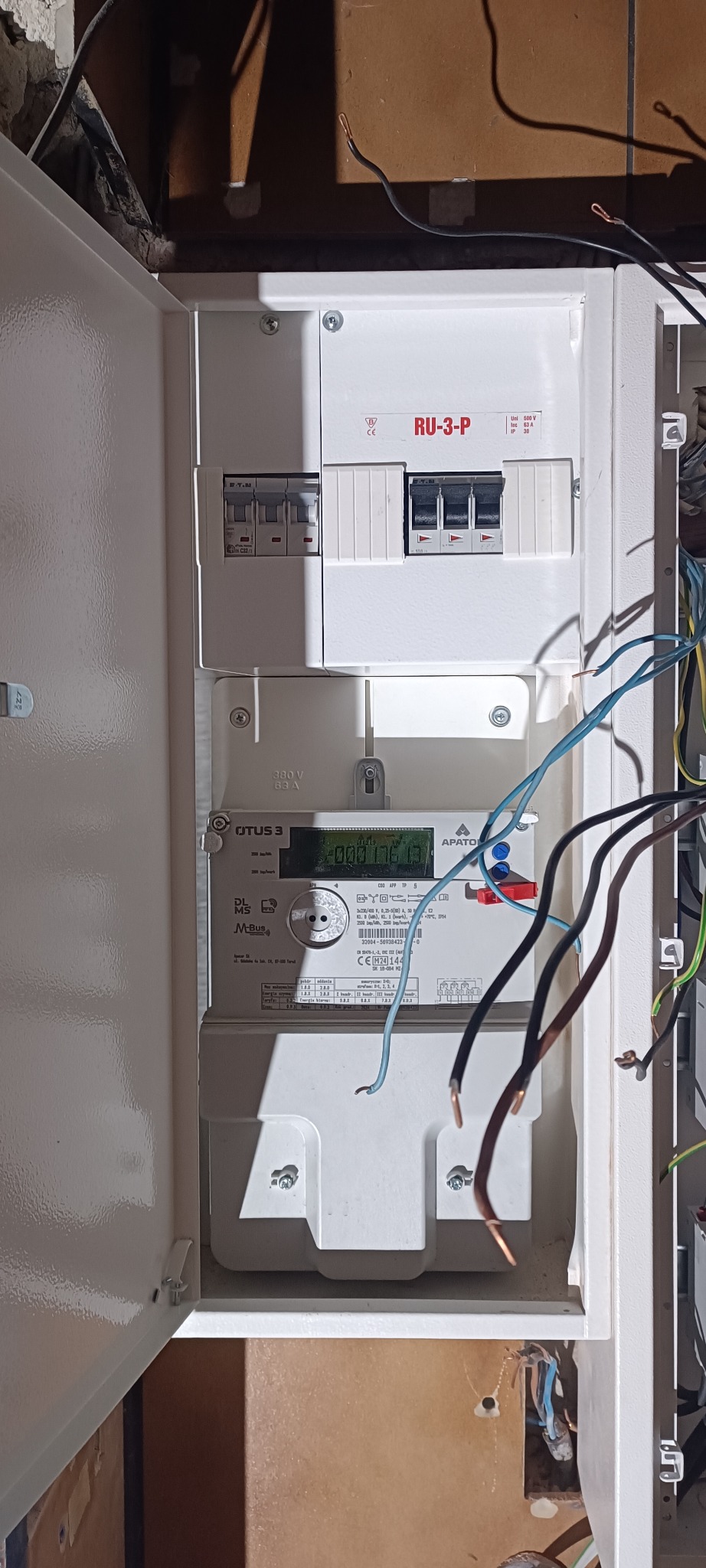 Otwarta skrzynka elektryczna z licznikiem i bezpiecznikami. Widoczne kable i elementy instalacji elektrycznej. Skupienie na szczegółach montażu.